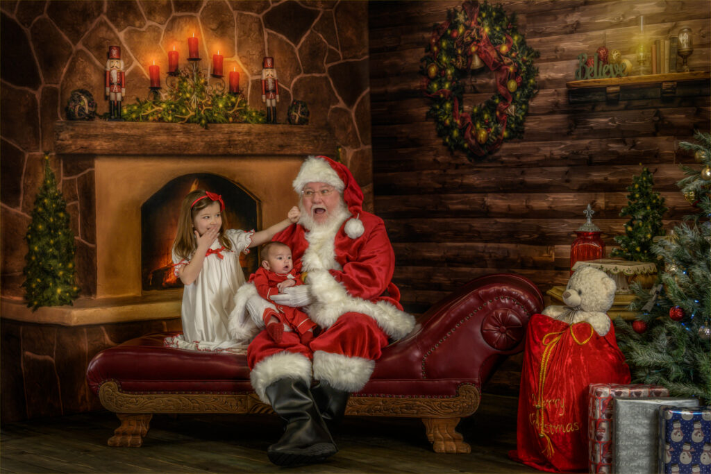 Little girl pulling Santa’s beard while holding her baby brother