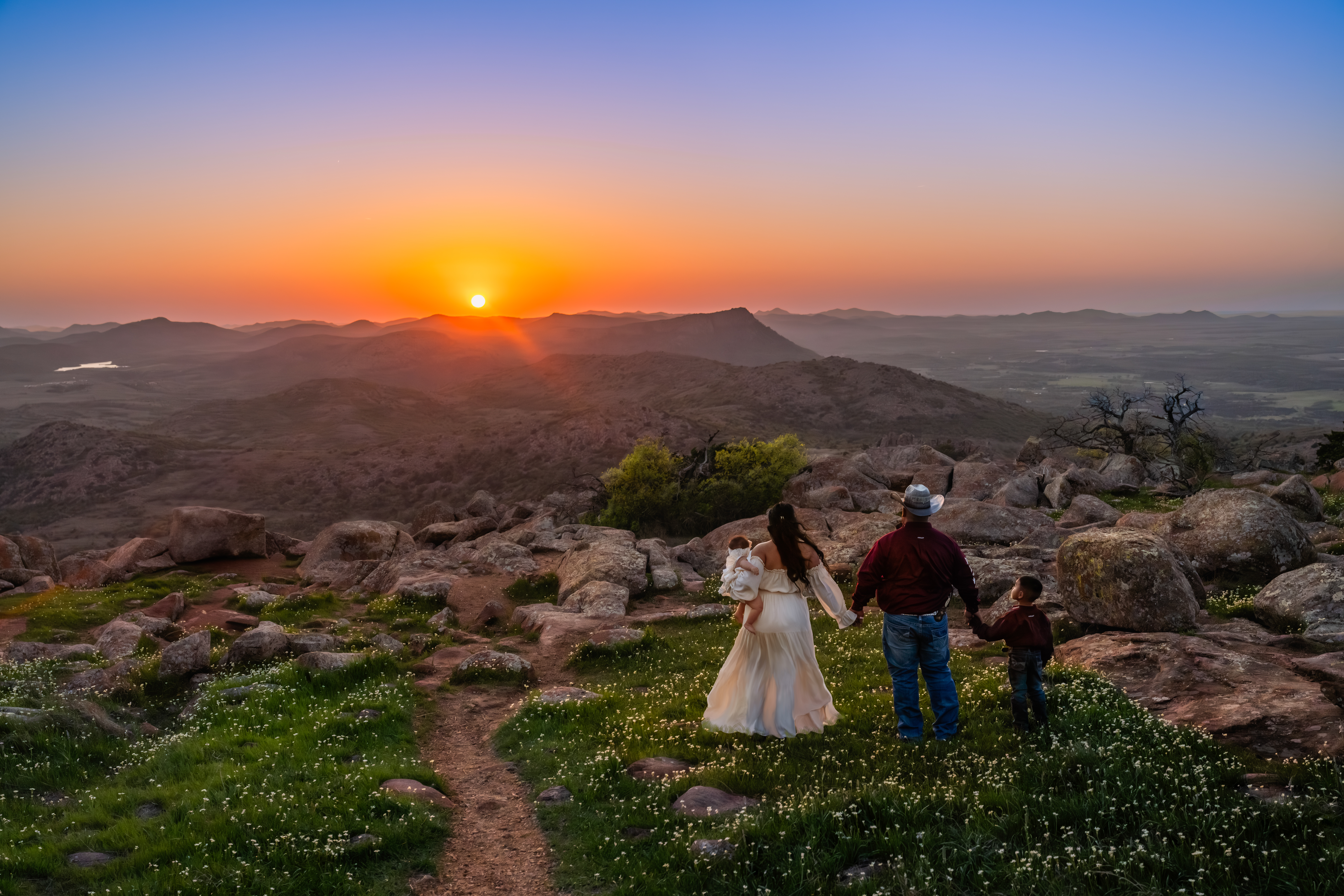 A young family of four stands together on top of the Wichita Mountains at sunset, bathed in golden and purple light. The mother’s dress flows in the breeze as she holds her baby, while the father and son stand beside her, all facing the breathtaking view.