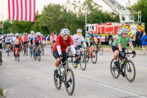 Riders in red and green jerseys leading a group under the American flag during the Hotter’N Hell Hundred 2025 start in Wichita Falls.