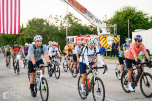 Group of cyclists in colorful jerseys riding under the American flag at the start of Hotter’N Hell Hundred 2025 in Wichita Falls