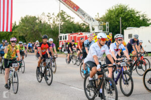 Rider in sunflower jersey waving while rolling out under the flag at race start.