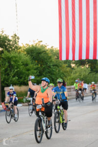 Rider taking a selfie under the American flag during Hotter’N Hell Hundred 2025 rollout in Wichita Falls.